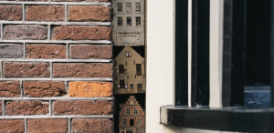 Banner image of the June 2023 issue of Words Without Border, featuring a brick wall and another wall with a window, in between which are toy-sized houses stacked vertically.